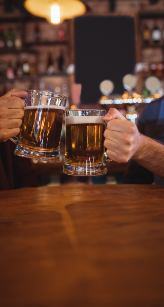 Two people clinking large beer mugs over a wooden table in a cozy bar with a warm, dimly lit atmosphere. The background is slightly blurred, showing shelves with various bottles and subtle light reflections, creating a lively but relaxed mood.