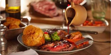 A plate of prime rib dinner is served with a Yorkshire pudding, roasted vegetables, and broccoli. A person is pouring gravy from a jug onto the meal. In the background, a platter of meat, a bowl of carrots, and a bottle of wine are visible.