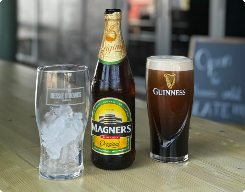 A bottle of Magners Original Irish Cider stands between an empty glass filled with ice on the left and a glass of Guinness beer on the right. The setup is on a wooden table, with blurred background elements including a chalkboard sign.