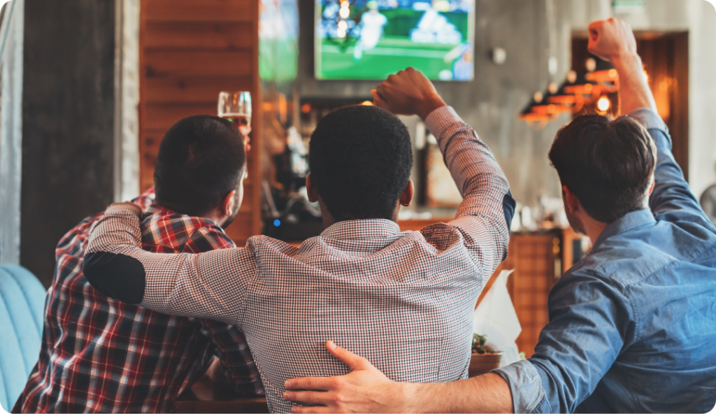 Three friends, with their backs facing the camera, sit side by side in a bar. They are cheering while watching a soccer match on a wall-mounted TV. One friend has his arm around another's shoulder, creating a lively and supportive atmosphere.